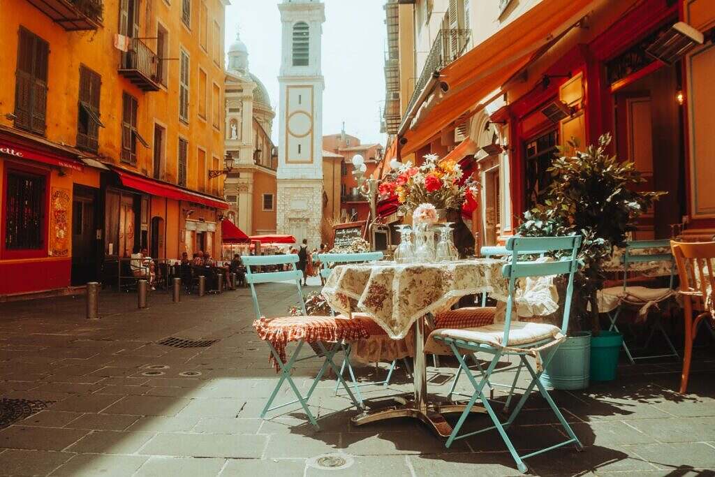 Utomhusserveringen på ett café i centrala Nice äldre delar, med gröna stolar runt ett bord placerade på den idylliska gågatan med ett kyrktorn i bakgrunden och vackra hus på vardera sida om gatan.