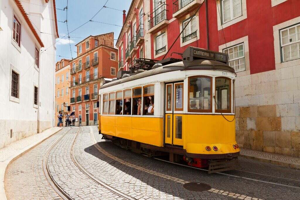 En gul, vintage spårvagn kör upp för en brant backe i Lissabon, Portugal utanför äldre, vackra bostadshus.