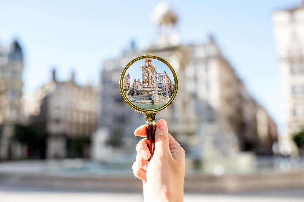 En hand håller ett förstoringsglas i handen framför en av Lyons berömda fontäner La Place des Jacobins.