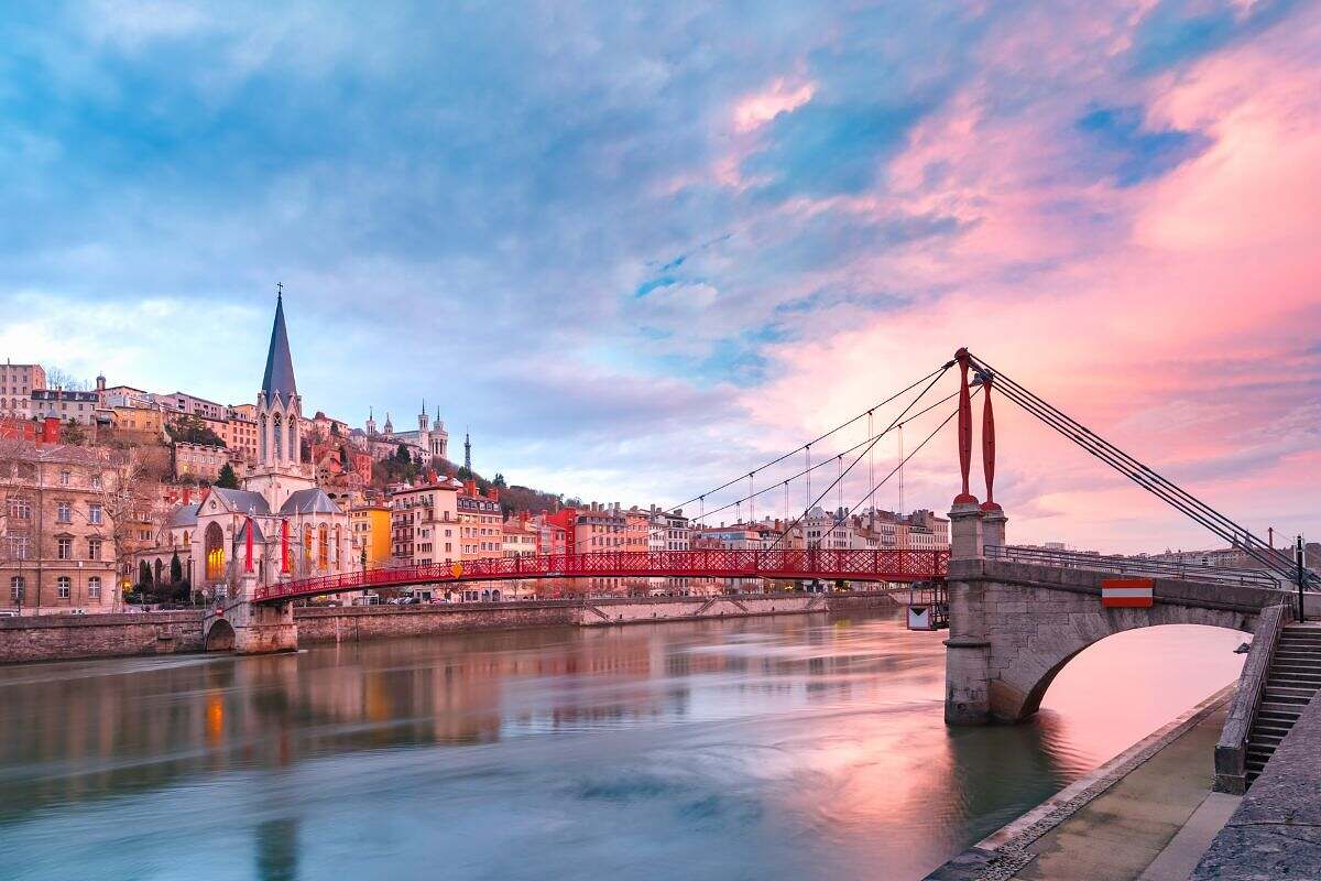 En röd gångbro i centrala Lyon, Frankrike med vackra äldre hus och en blå och rosa himmel i skymningen.