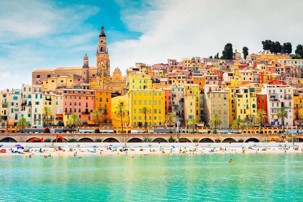 Det turkosgröna havet utanför Nice, Frankrike med vackra gama hus på en kulle och ett kyrktorn bakom turister som badar.