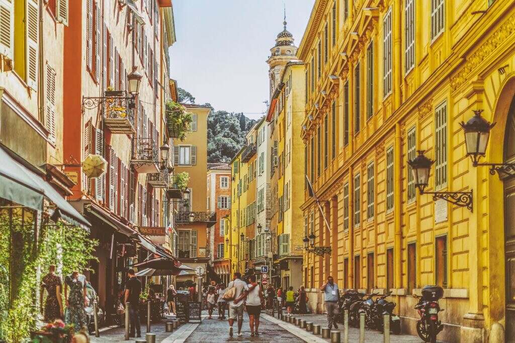Vacker gammal gågata i Nice, Frankrike med människor som promenerar på en gågata kantad med hus från 1800-talet och ett kyrktorn i bakgrunden.