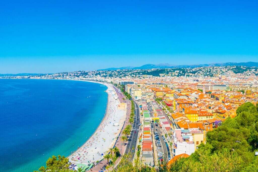 En scenisk vy över kusten utanför staden Nice i Frankrike med det blåa havet till vänster och stadshusen och berg till höger.