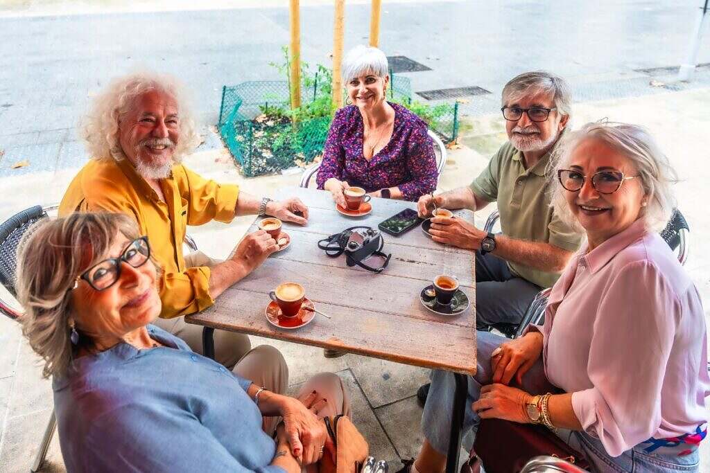 Fem leende pensionärer dricker kaffe tillsammans utomhus.