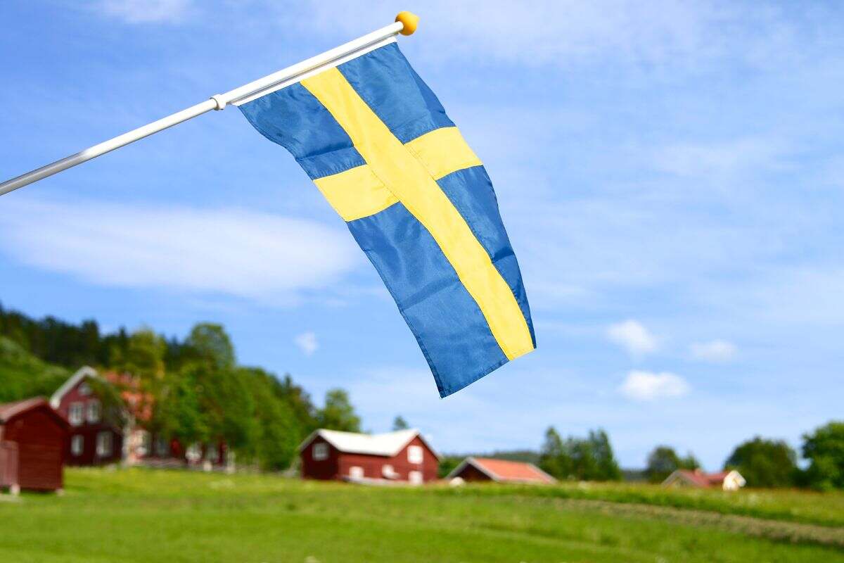 En svensk flagga vajar med röda hus och grönska i bakgrunden.