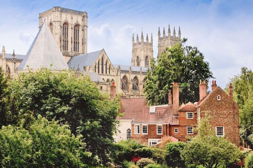 Idylliskt bild av York Minster Cathedral i York, UK med små tegelstenshus utanför och lummiga träd.